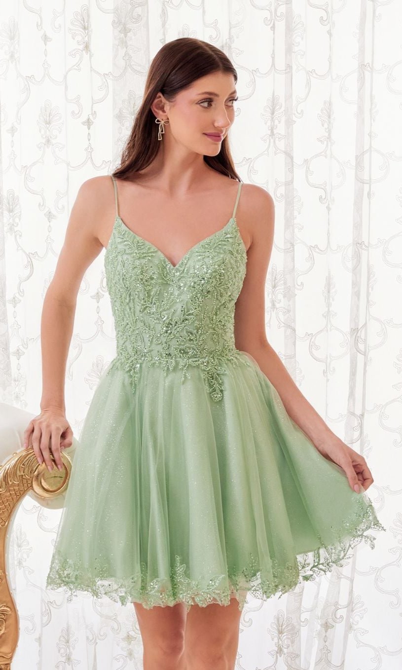 A woman wears the Short Homecoming Dress CD0213 with embroidery, featuring light green lace and tulle, sleeveless and short, as she stands beside a chair against a sheer, patterned curtain.